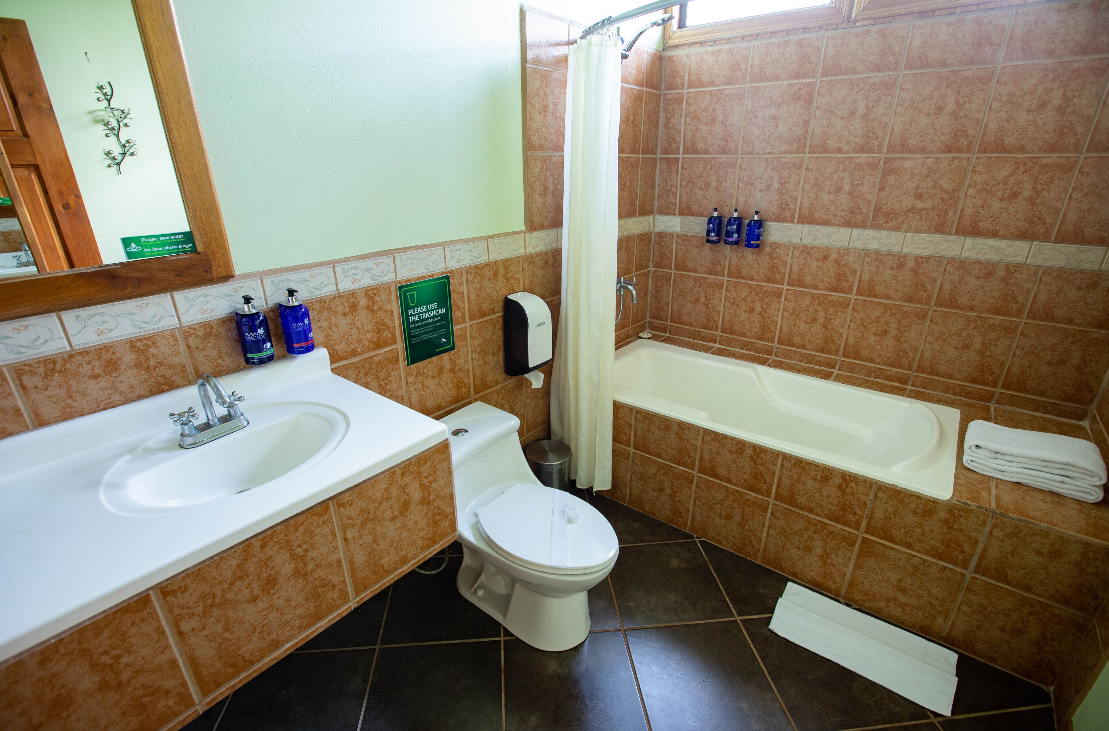 A bathroom with a sink, toilet, and bathtub. There are toiletries on the counter and a sign instructing to use the trash can.
