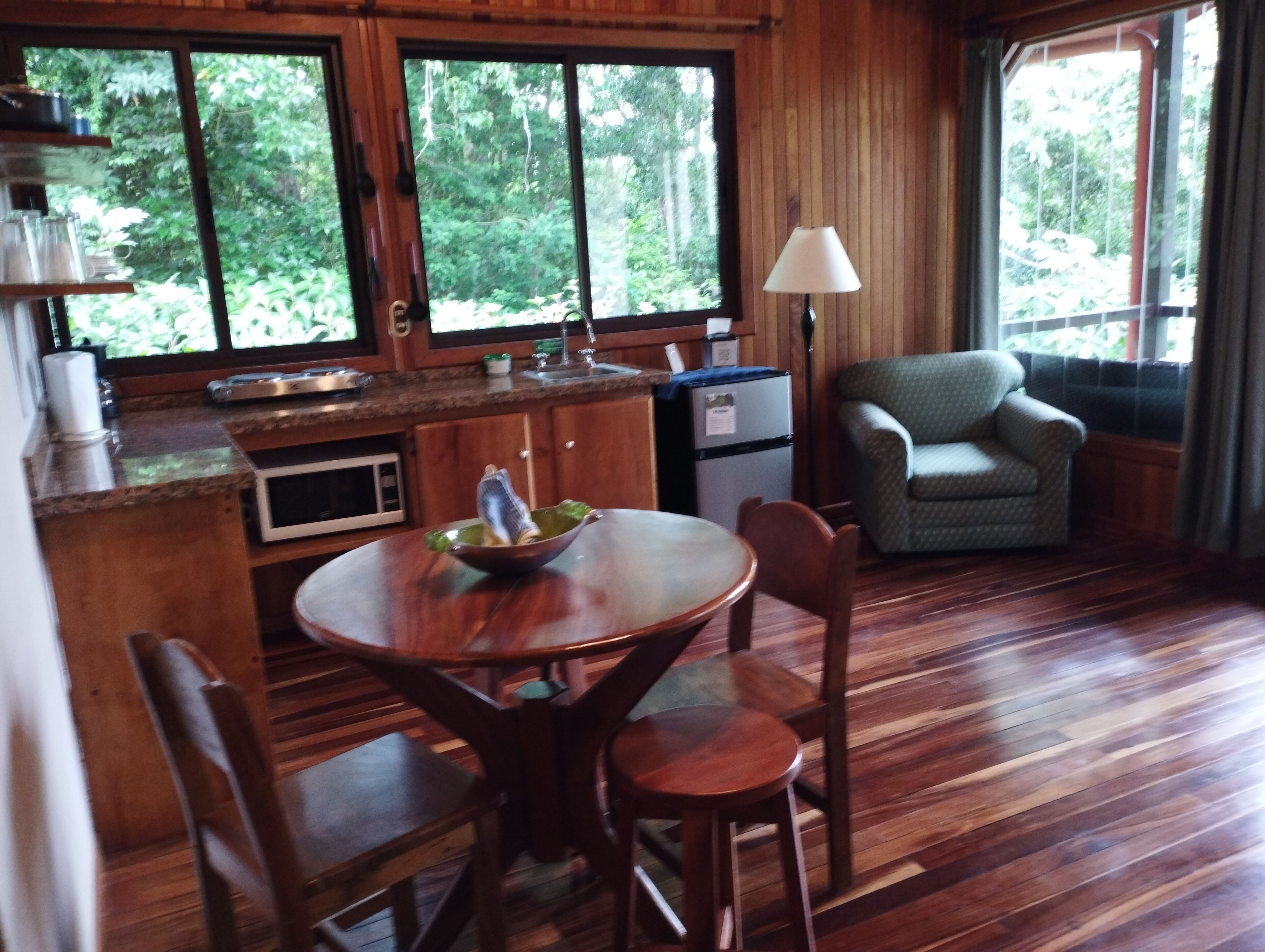 A wooden cabin interior with a dining table, chairs, a kitchen area, and a view of the forest through large windows.
