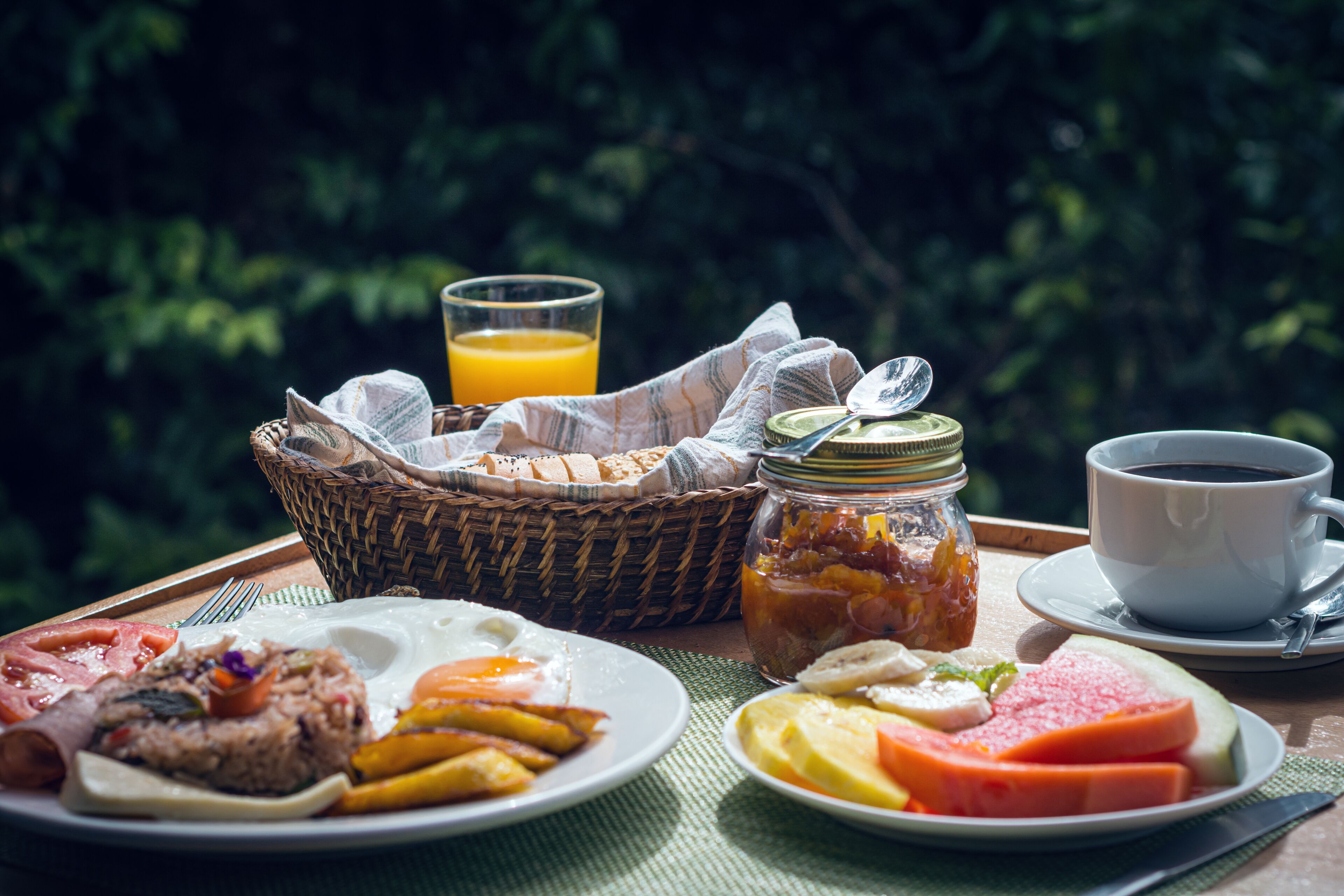 A breakfast setting with a basket, a glass of orange juice, a cup of coffee, and plates of food including eggs, toast, and fruit.