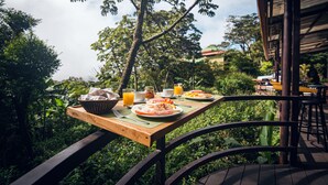 Mesa de madeira posta com itens de café da manh ã, cercada por vegetação exuberante e vista para um edifício ao fundo.