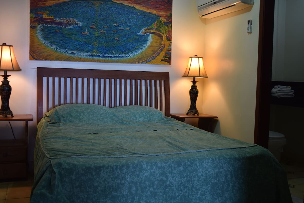 Ein Schlafzimmer mit einem Bett, zwei Nachttischlampen, einem Kopfteil, einem Gemälde mit Küstenlandschaft und einem durchgehenden Türspalt sichtbaren Badezimmer.