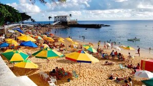 Een strand met kleurrijke parasols, mensen die zonnebaden, en een gebouw op de achtergrond.