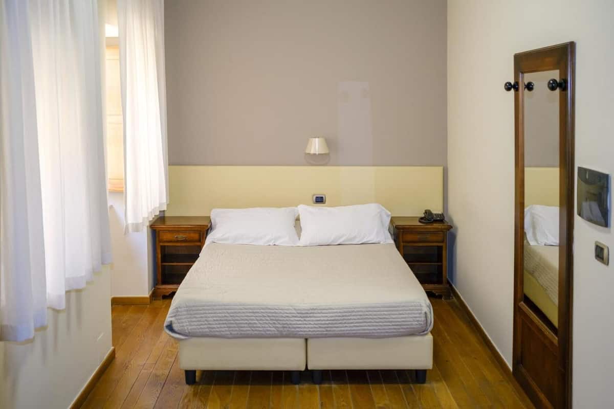 A neatly made bed with white linens in a room with wooden flooring, a bedside table with a lamp, and a mirror on the wall.