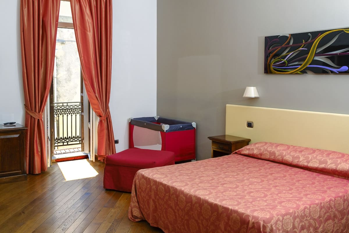 A hotel room with a wooden floor, a bed with a red bedspread, a small red chair, a nightstand, a lamp, and a painting on the wall.