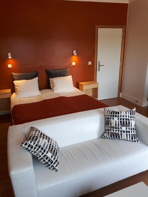 A hotel room with a bed, bedside tables, a white sofa with patterned cushions, and a door.