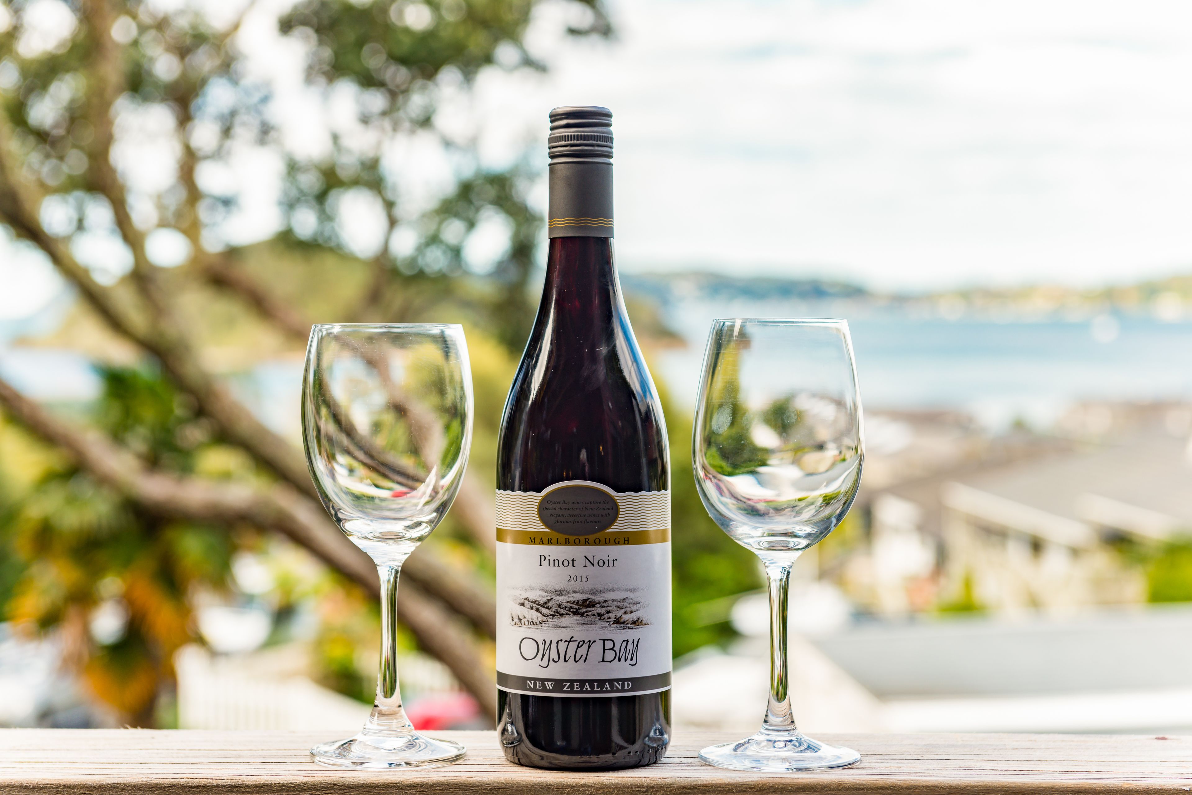 Eine Flasche Pinot Noir Wein mit zwei leeren Gläsern auf einem Holztisch, vor verschwommenem Hintergrund im Freien.