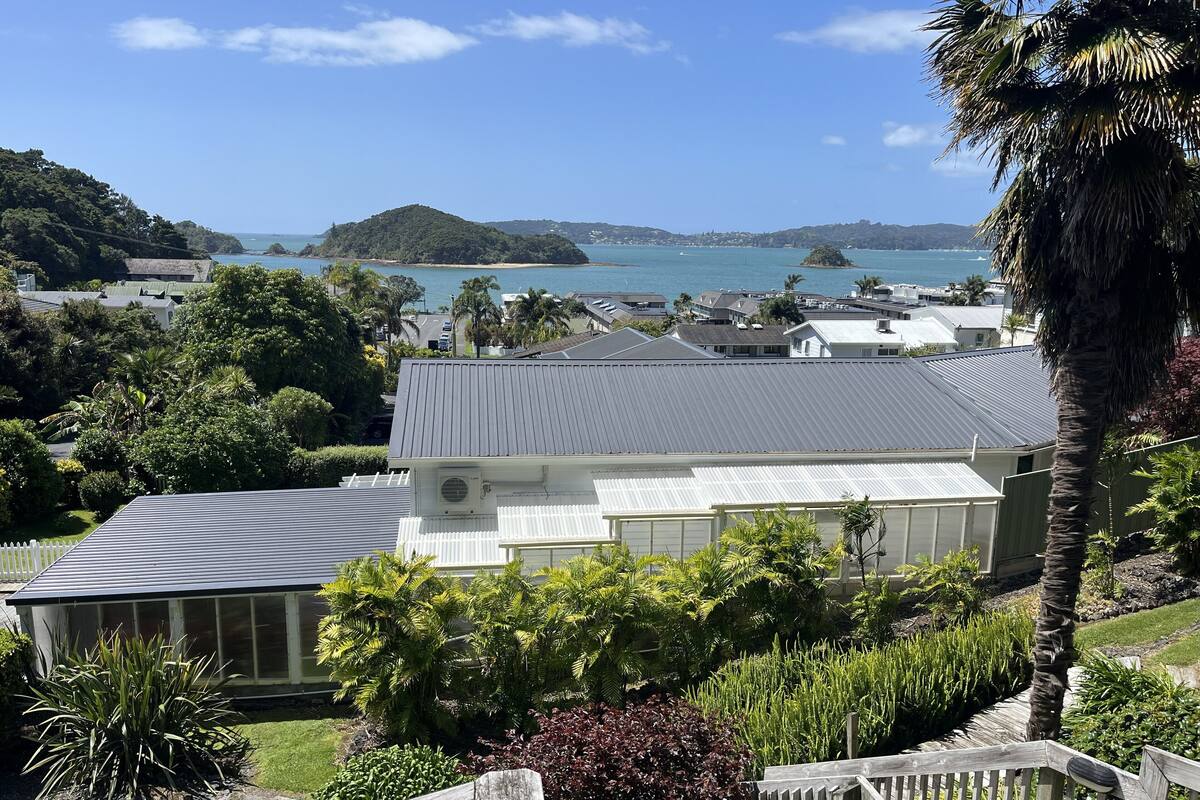A residential area with a house featuring a grey roof and white walls, surrounded by lush greenery and a view of the sea.