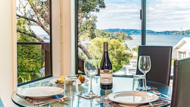 Una mesa de comedor con una botella de vino, platos y cubiertos, frente a una vista panorámica de un lago y colinas.