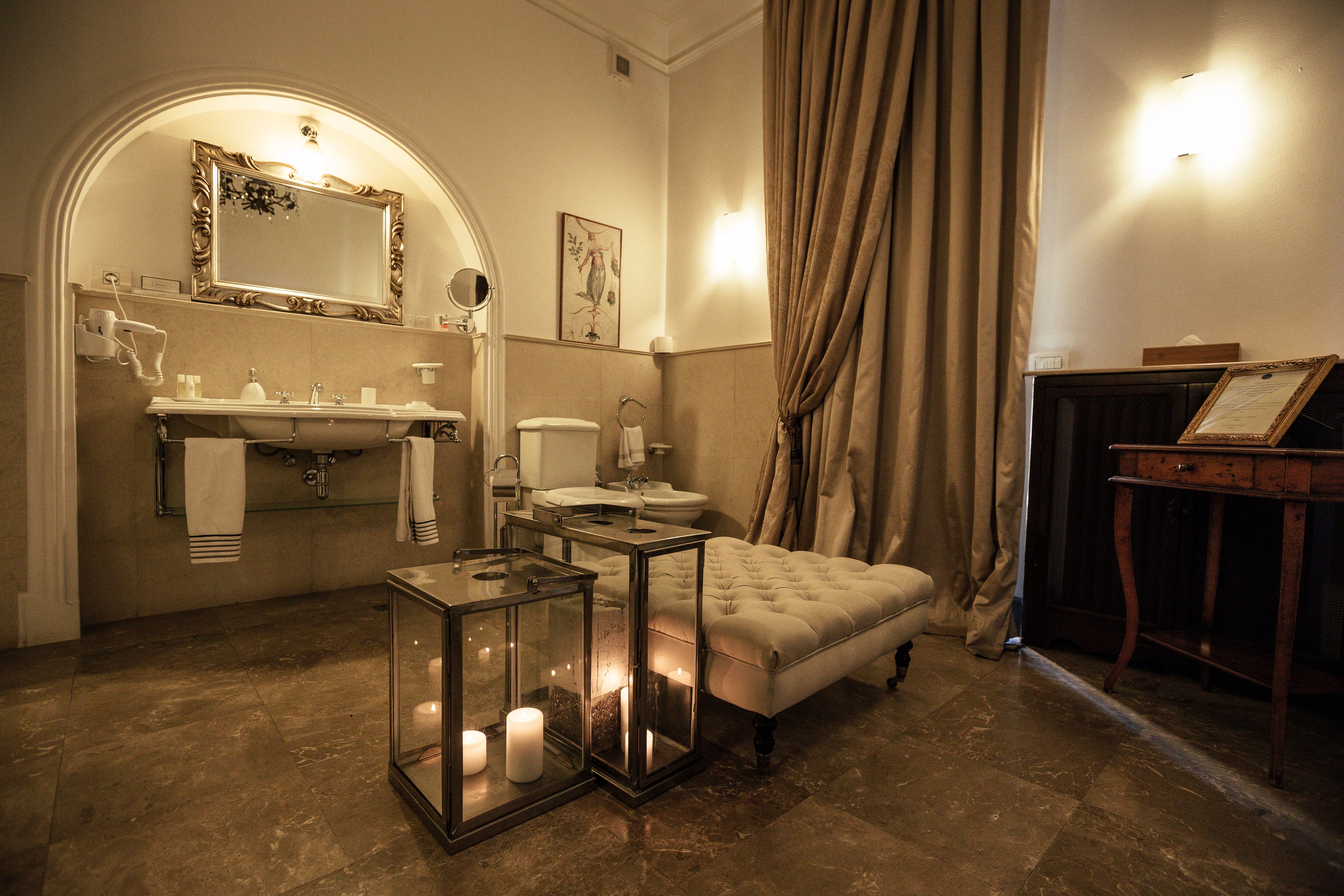A bathroom with a vanity, sink, toilet, and a cushioned bench. There are candles in glass lanterns on the floor.