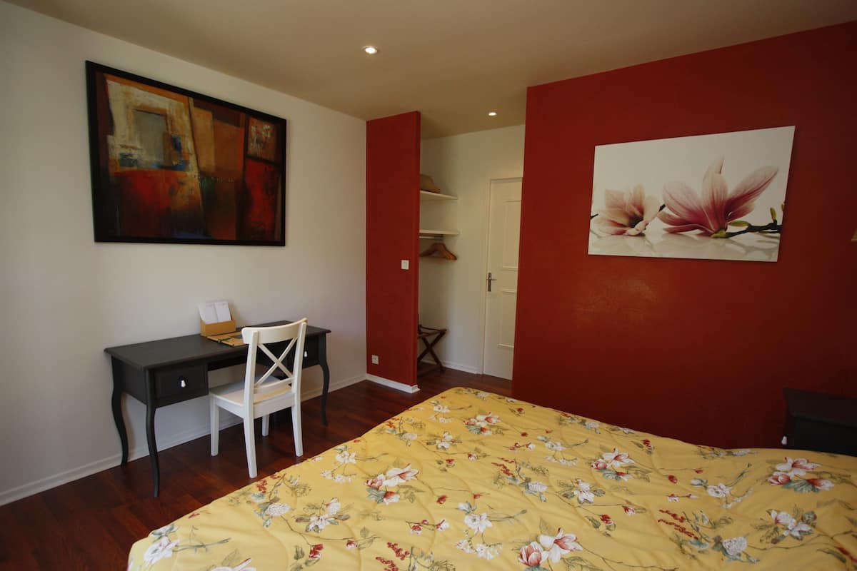 A bedroom with a bed, a desk, and a chair. There are two paintings on the walls.
