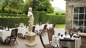 Un espace de restauration en plein air avec des tables dressées pour un repas, une statue et un mur de pierre agrémenté d’une fenêtre.