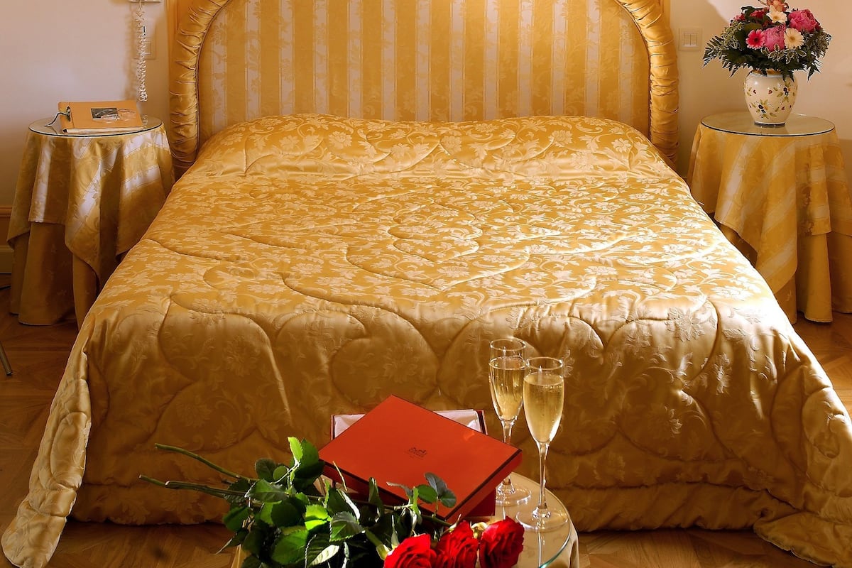 A hotel room with a large bed, a chandelier, a bedside table with a vase of flowers, and a small table with a bouquet of roses and a red box.