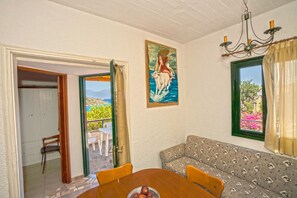 A dining area with a table, chairs, and a bowl of fruit. There is a painting on the wall and a window with a view of greenery.