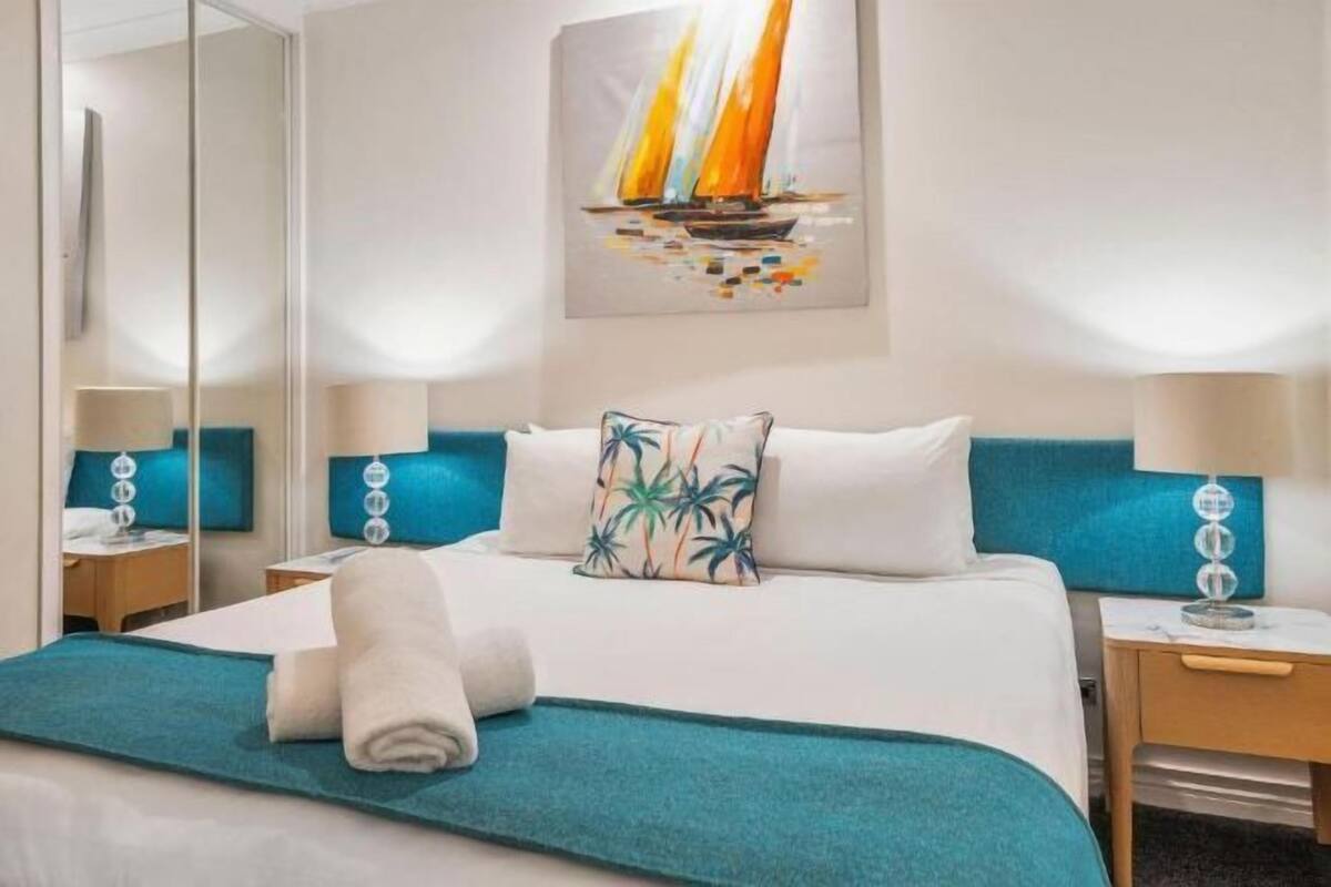 A hotel room with a bed, a nightstand, a mirror, a painting of a sailboat, and a teal throw blanket.