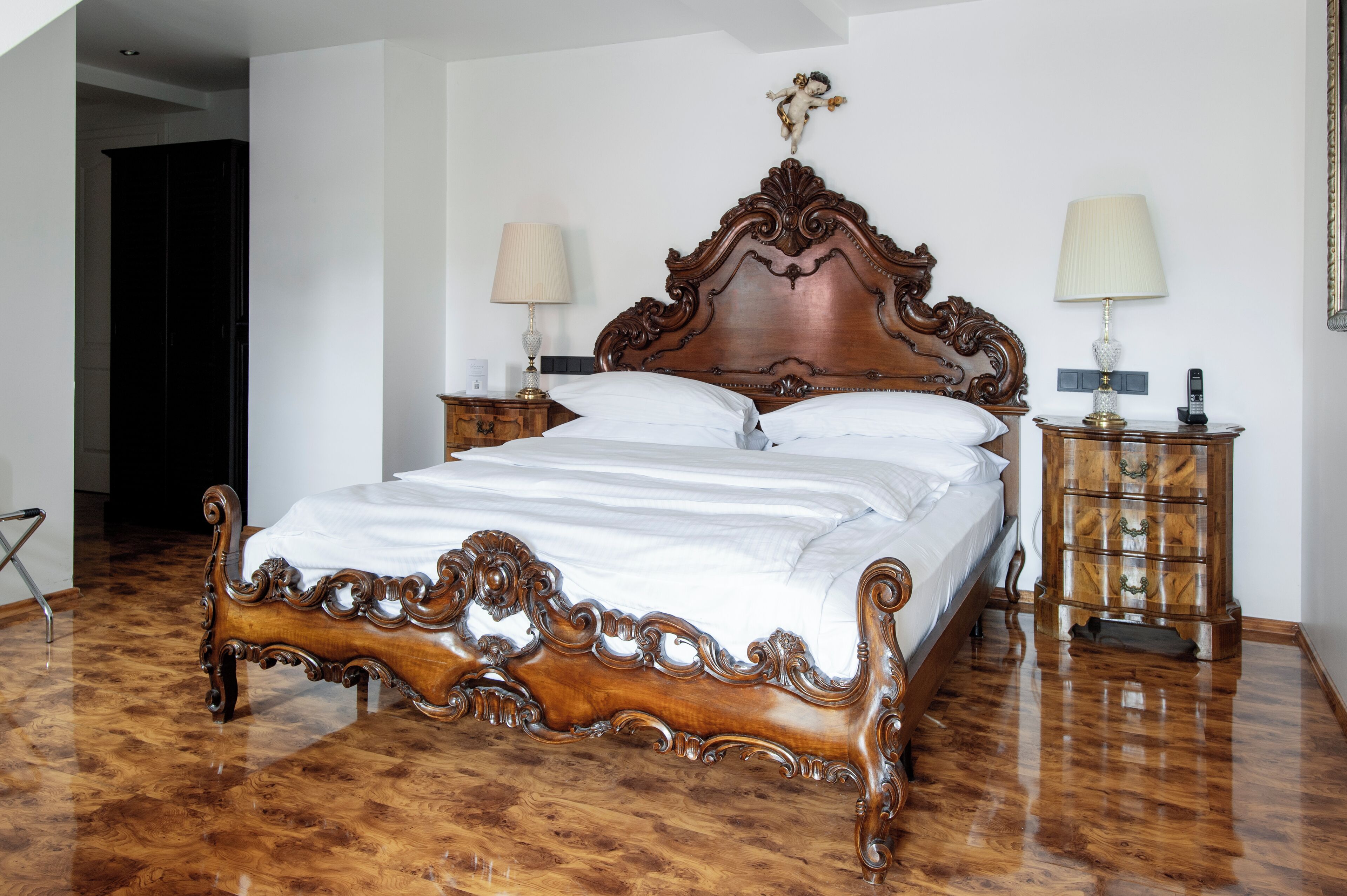 Uma cama de madeira com detalhes entalhados, duas mesas de cabeceira com abajures e um espelho à direita.