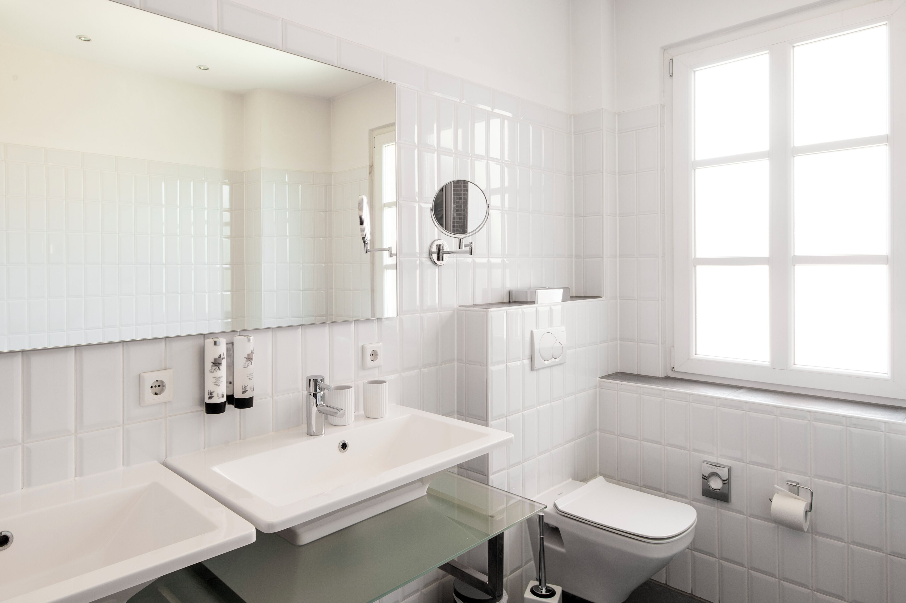 Une salle de bain moderne avec des carreaux blancs, une étagère en verre, un lavabo, des toilettes, un miroir et une fenêtre.