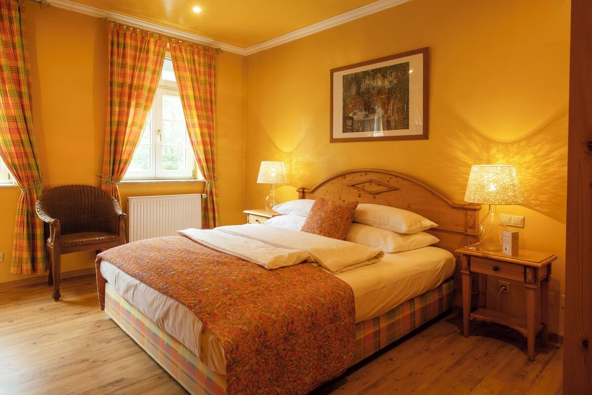 A bedroom with a bed, two bedside tables with lamps, a chair, a window with curtains, and a painting on the wall.
