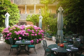 An outdoor patio with round tables and chairs, surrounded by lush greenery and pink flowers. - Werners Boutique Hotel (Fuerth)