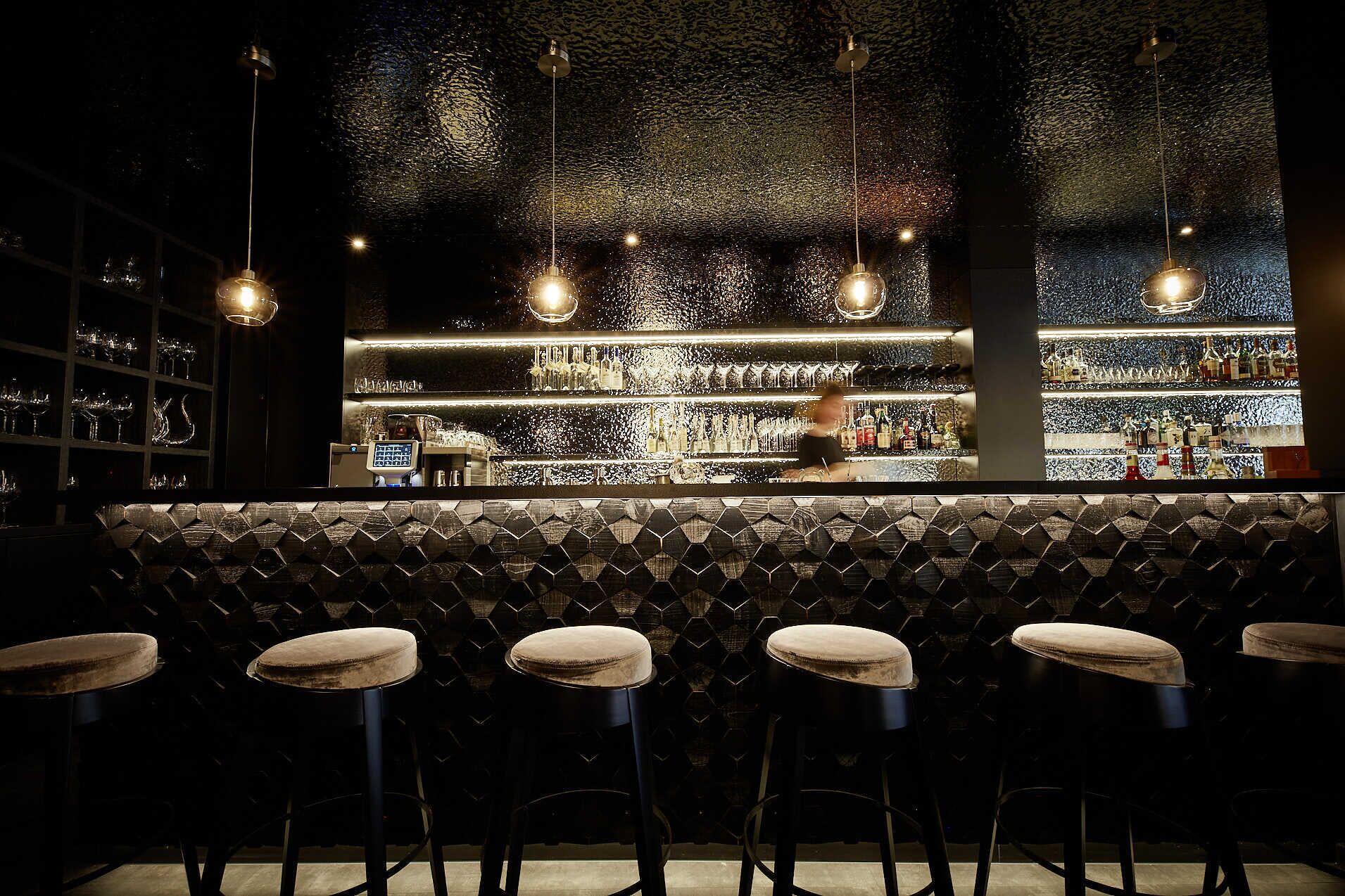 Un bar con pared de diseño hexagonal, taburetes y lámparas colgantes.
