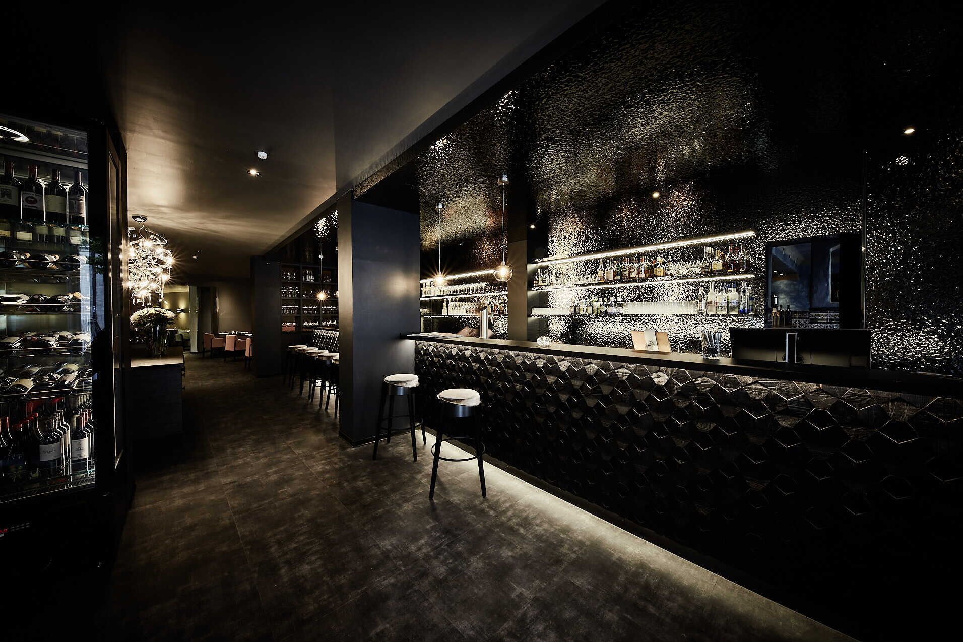 A dimly lit bar area with a wine rack, bar stools, and a patterned wall.
