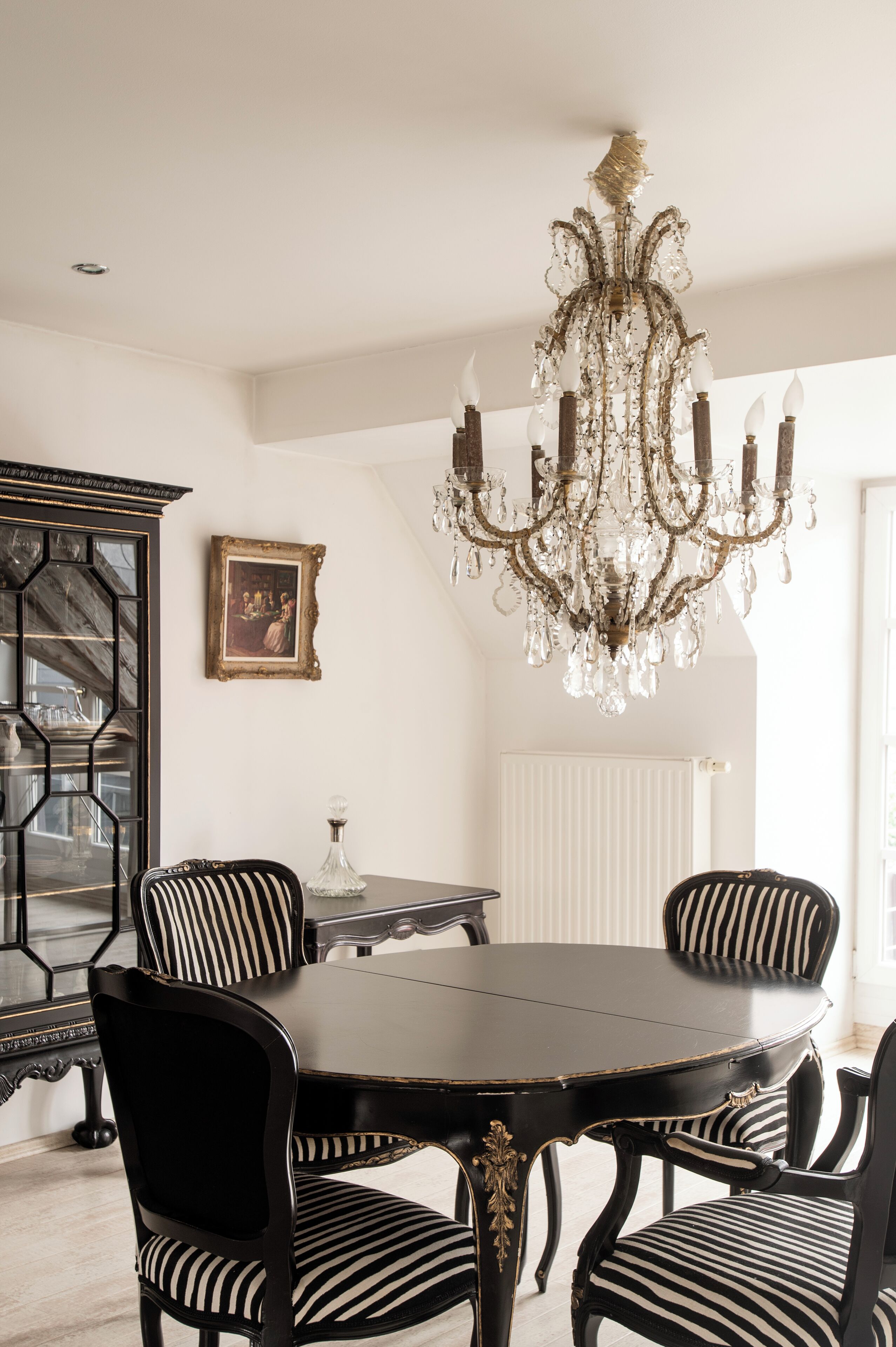 A dining area with a round table, four chairs, a chandelier, a cabinet, and a framed picture on the wall.
