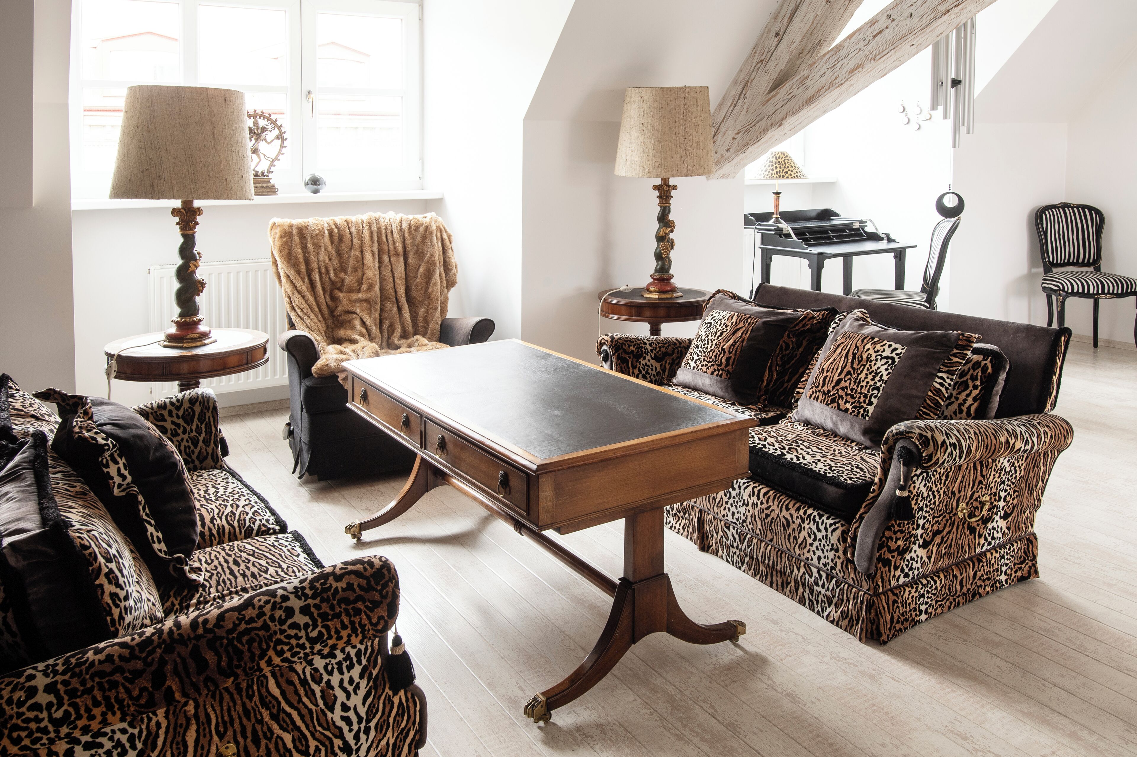 Een woonkamer met een houten salontafel, sofa's met luipaardprint en fauteuils. Twee lampen staan op bijzettafeltjes, en er is een piano op de achtergrond.