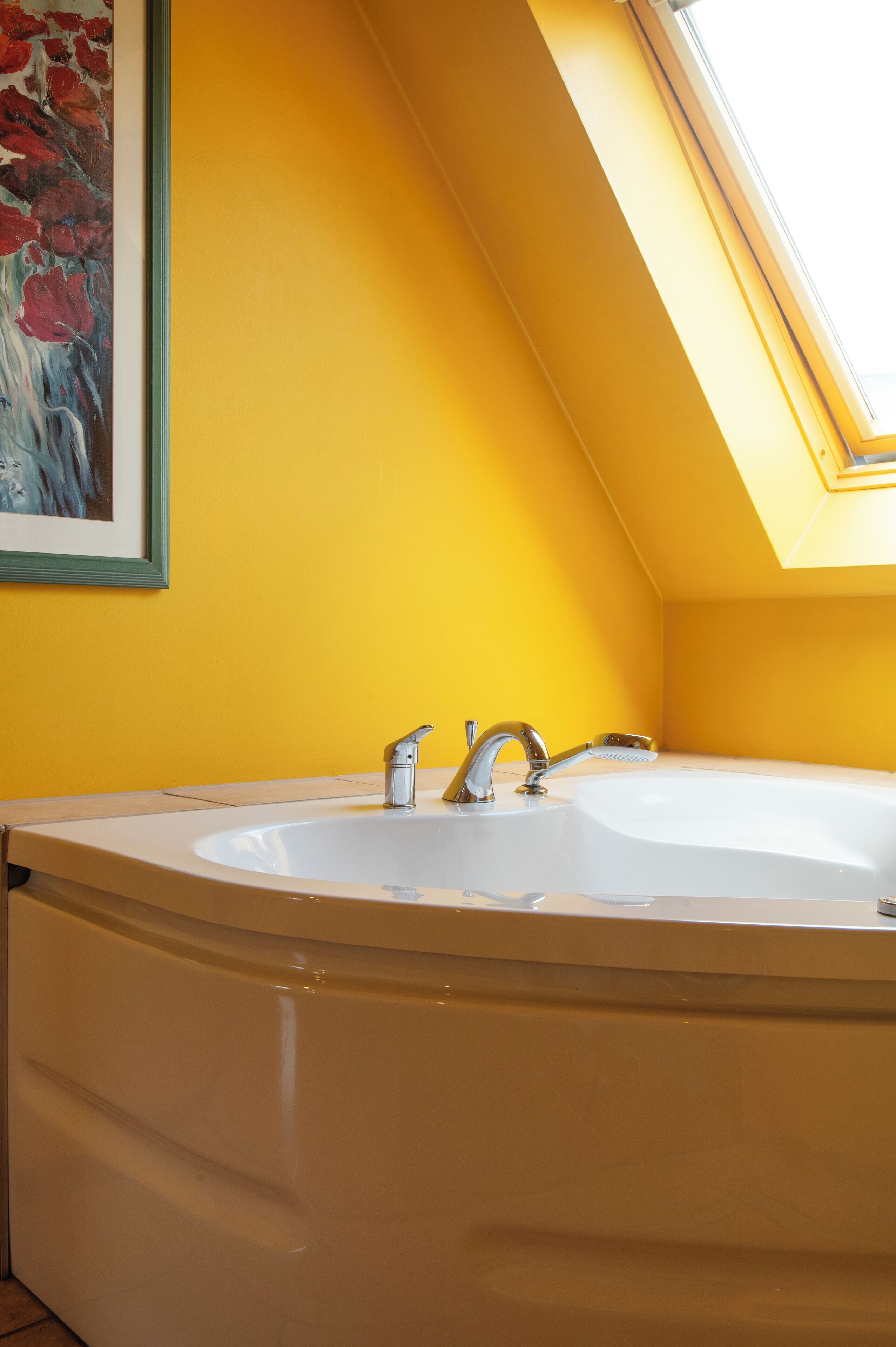 A bathroom with a large bathtub, a skylight, and a painting on the wall.