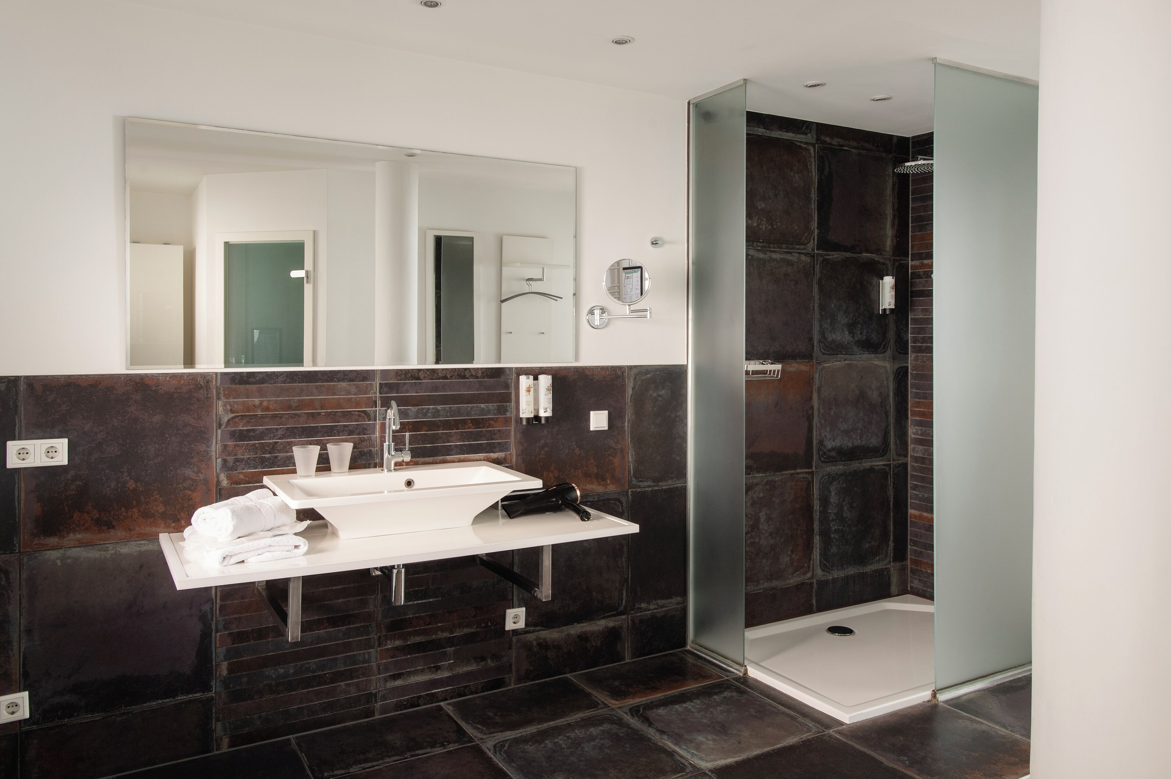 Un baño moderno con tocador blanco, paredes con azulejos oscuros, cabina de ducha de vidrio y un espejo grande.