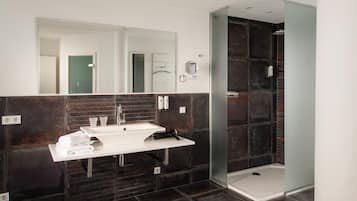 Un baño moderno con tocador blanco, paredes con azulejos oscuros, cabina de ducha de vidrio y un espejo grande.