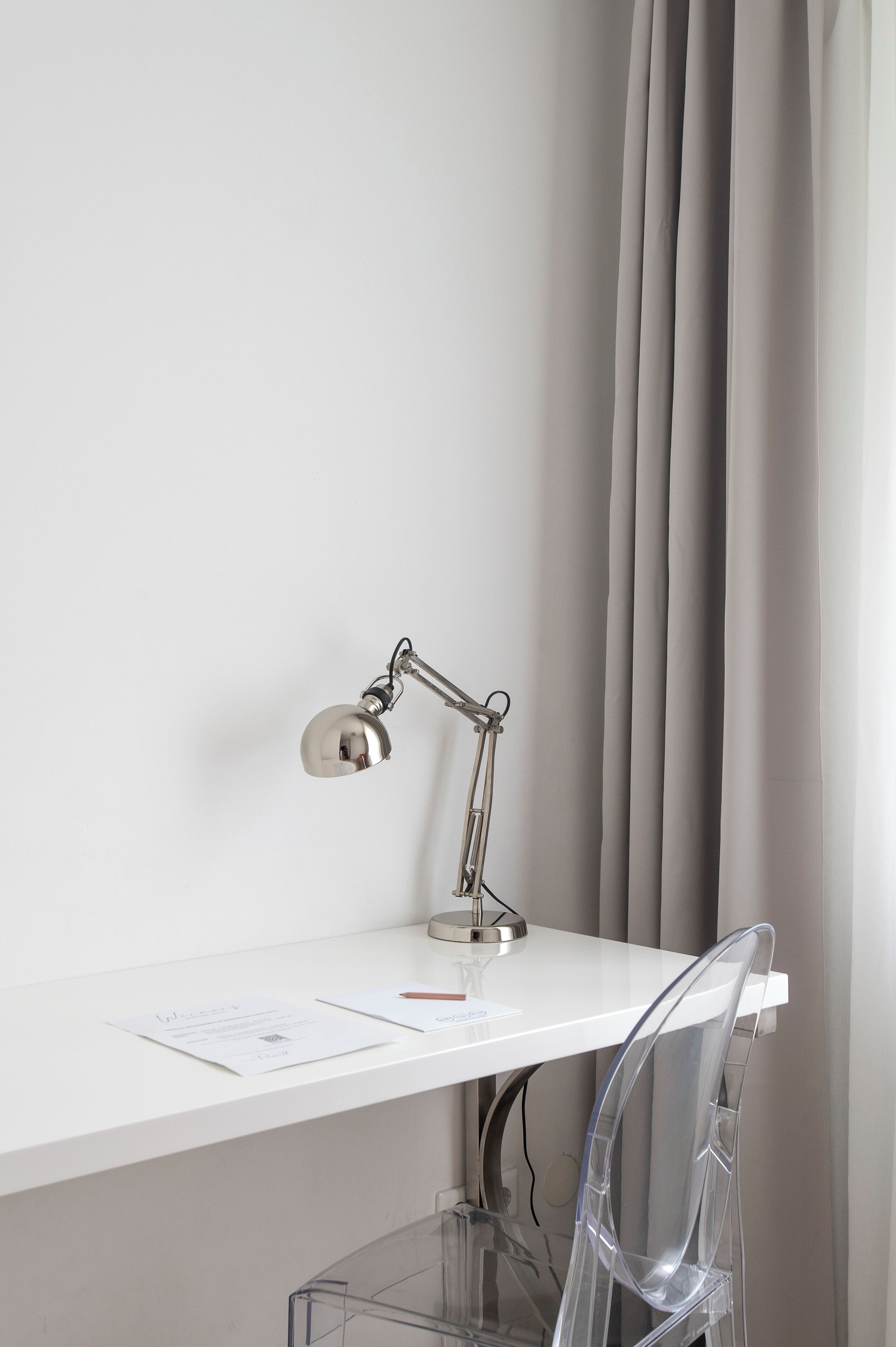 A white desk with a clear chair, a metallic desk lamp, and a curtain in the background.