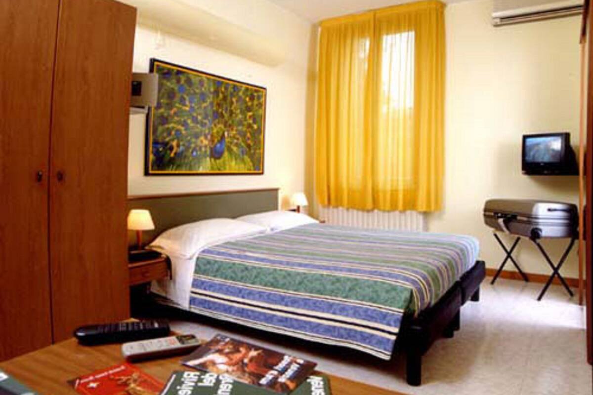 A hotel room with a bed, a desk, a television, a painting, and a wardrobe.