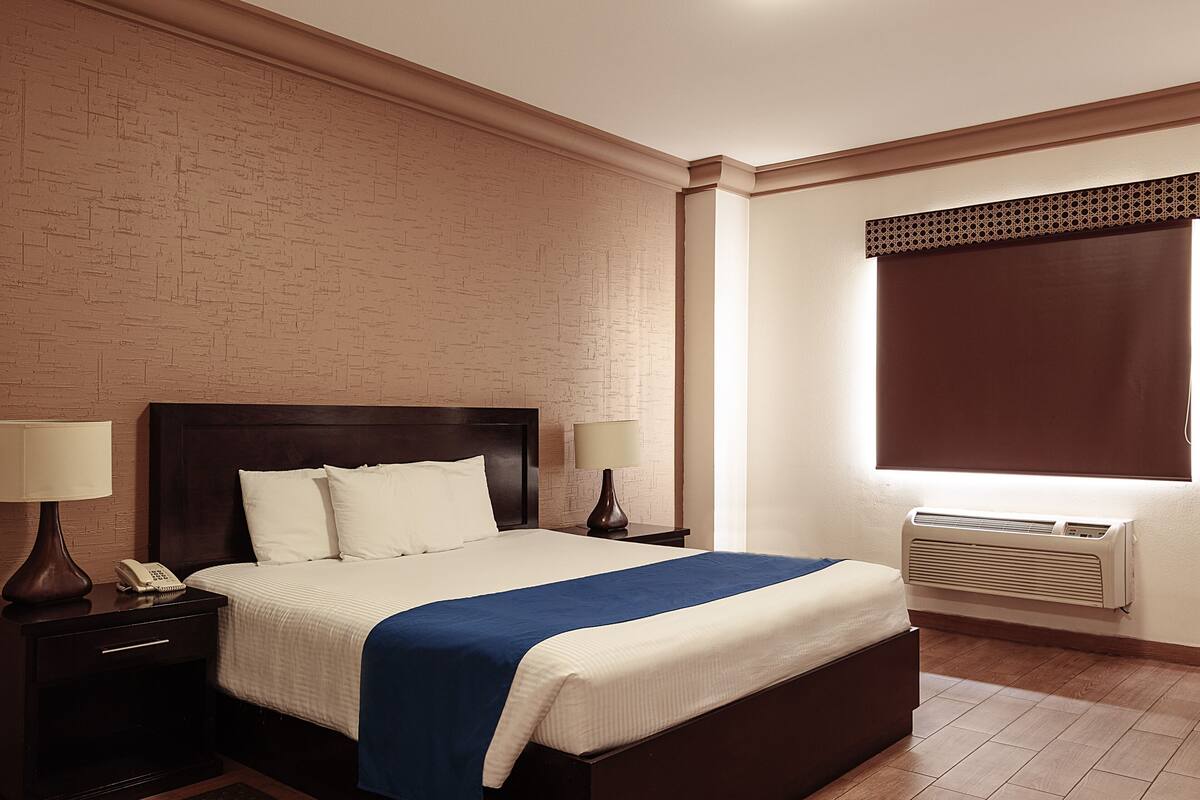 Hotellihuone, jossa on suuri sänky, kaksi yöpöytää valaisimineen, seinään asennettu ilmastointilaite ja ikkuna, jossa on sälekaihtimet.