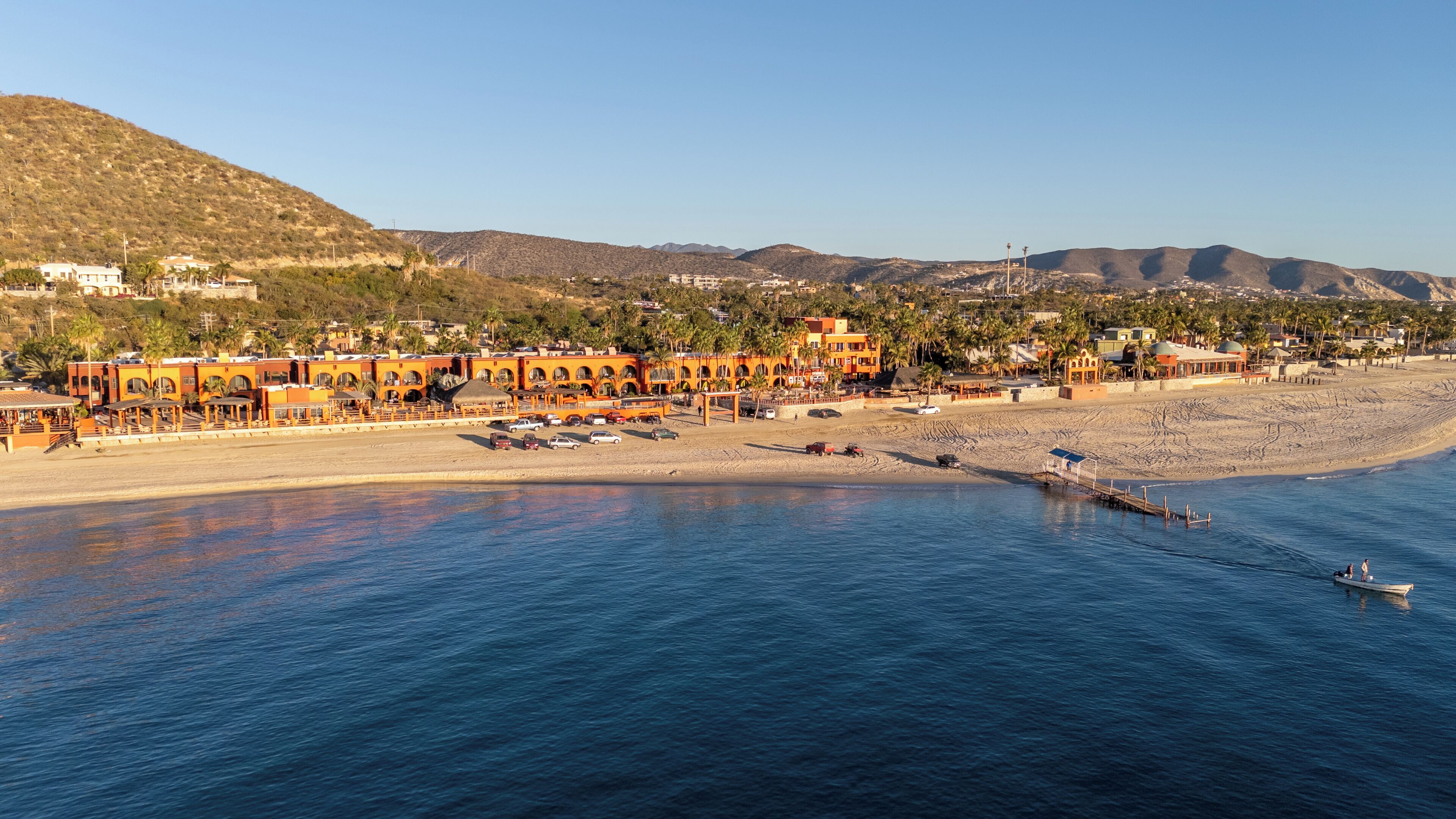 Un hotel costero con varias edificaciones, una playa y montañas al fondo.