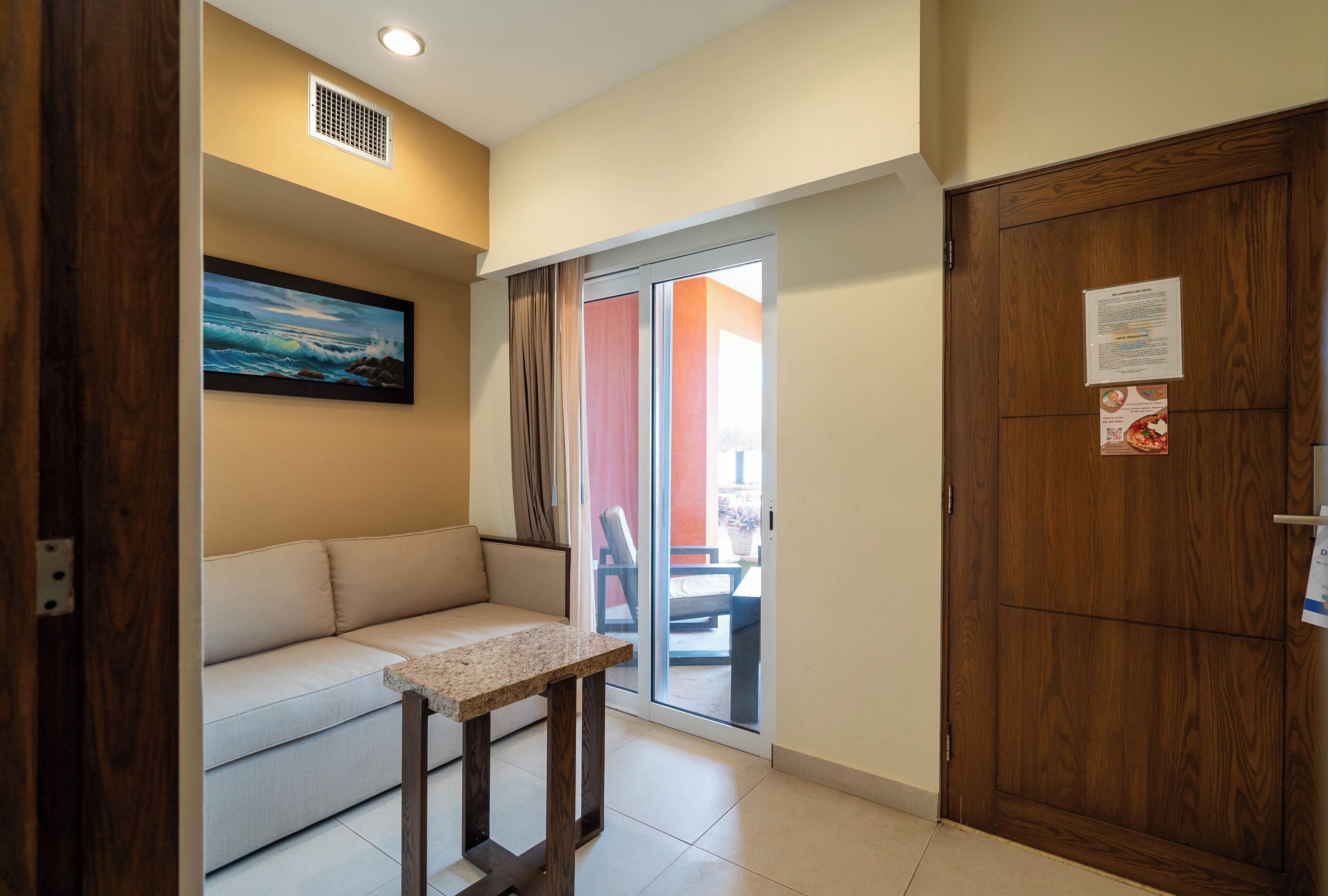 A room with a wooden door, a sliding glass door leading to a balcony, a beige sofa, and a small table.