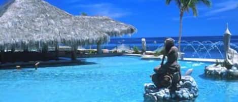 Une piscine avec une personne assise sur un rocher, des palmiers et une structure surmontée d’un toit de chaume.