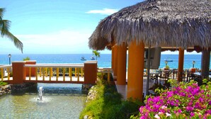 Un quiosco con techo de paja junto a una piscina, rodeado de flores coloridas y con vistas al mar azul y transparente.