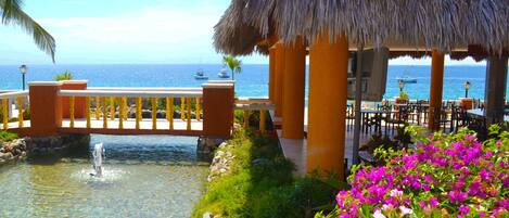 Un quiosco con techo de paja junto a una piscina, rodeado de flores coloridas y con vistas al mar azul y transparente.