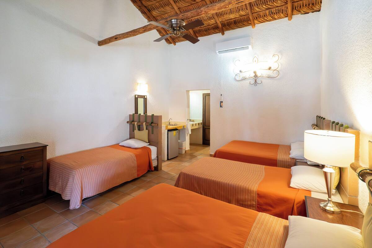 Une chambre d’hôtel avec trois lits, un plafond en bois, un petit bureau, un miroir et une table de chevet avec une lampe.