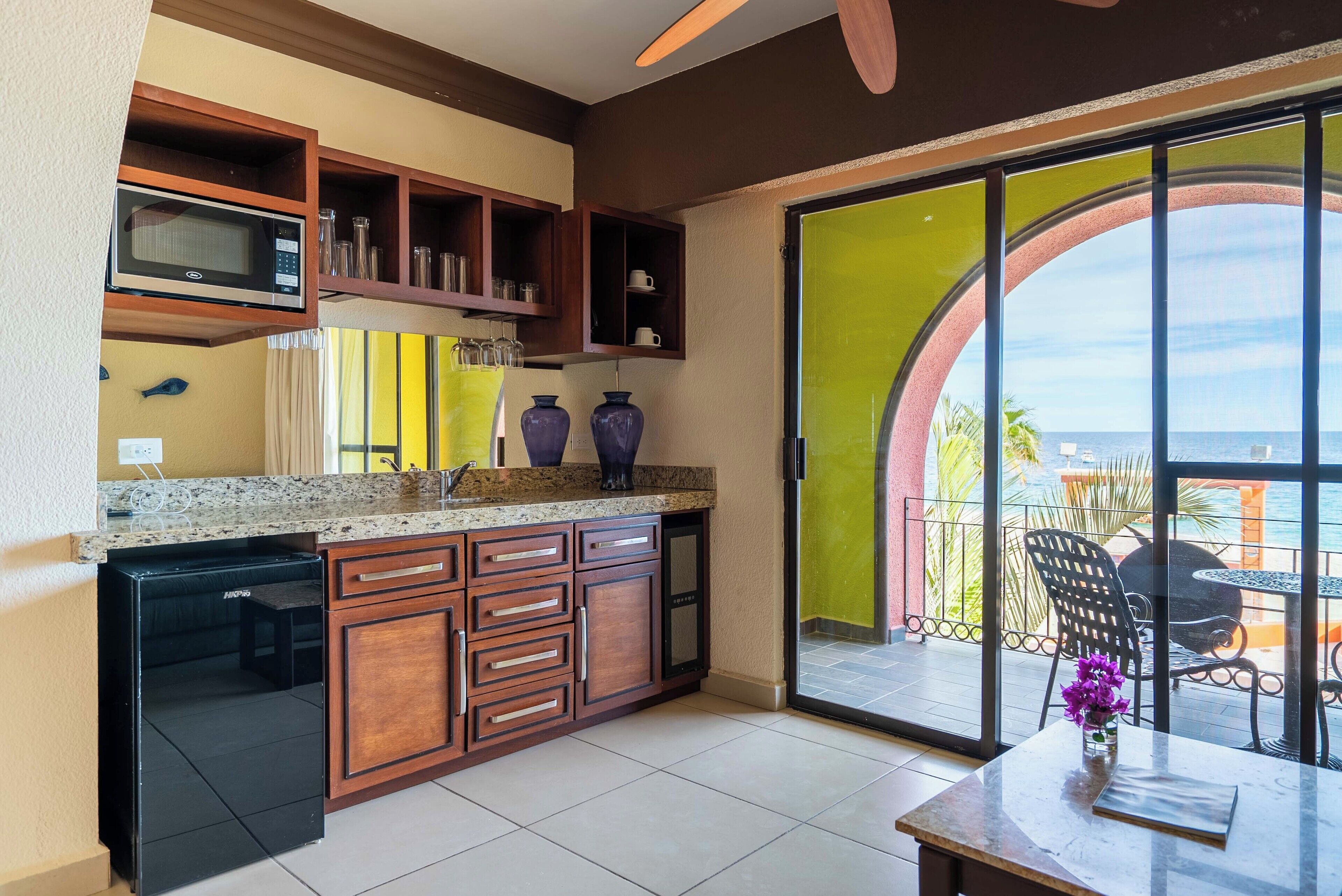 A kitchen with wooden cabinets, a microwave, and a refrigerator. There is a dining area with a table and chairs, and a view of the ocean through large windows.