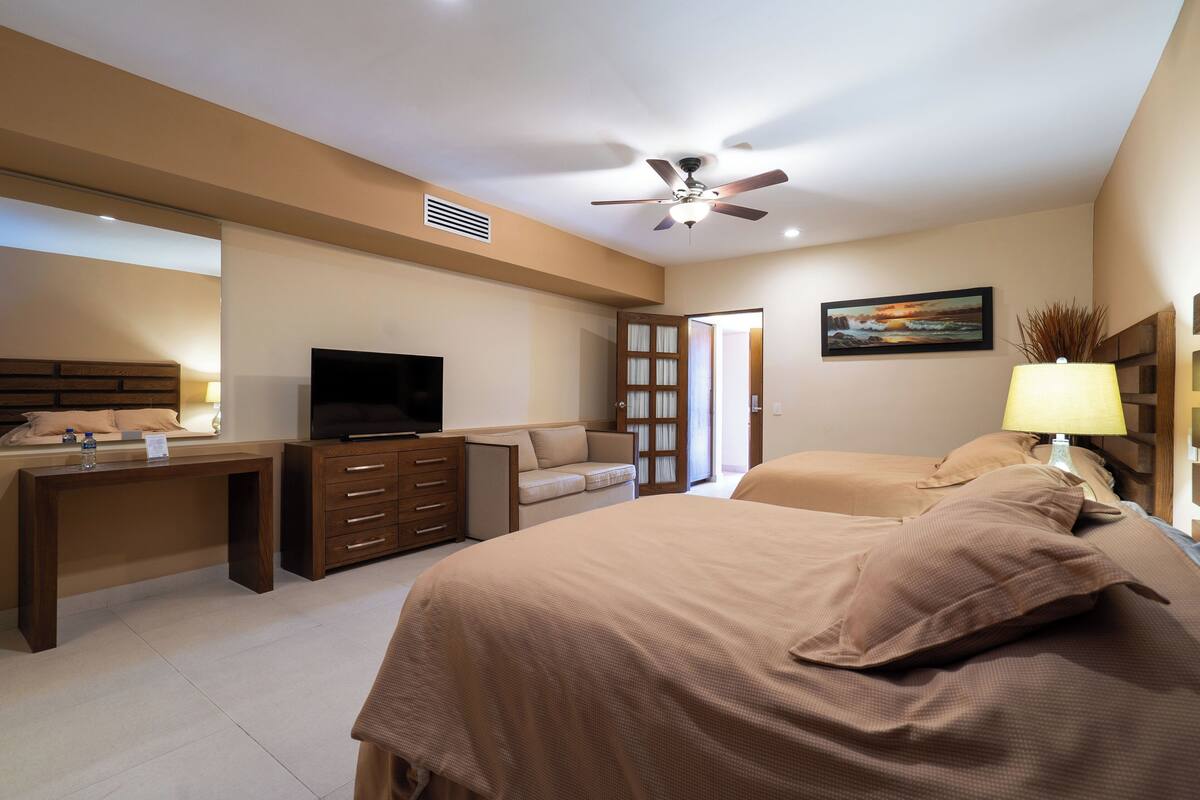 Une chambre à coucher comprenant un lit, un bureau, une télévision, un ventilateur de plafond et un tableau accroché au mur.