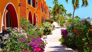 Un hotel con fachada roja, ventanas arqueadas y un camino bordeado de flores coloridas y palmeras.