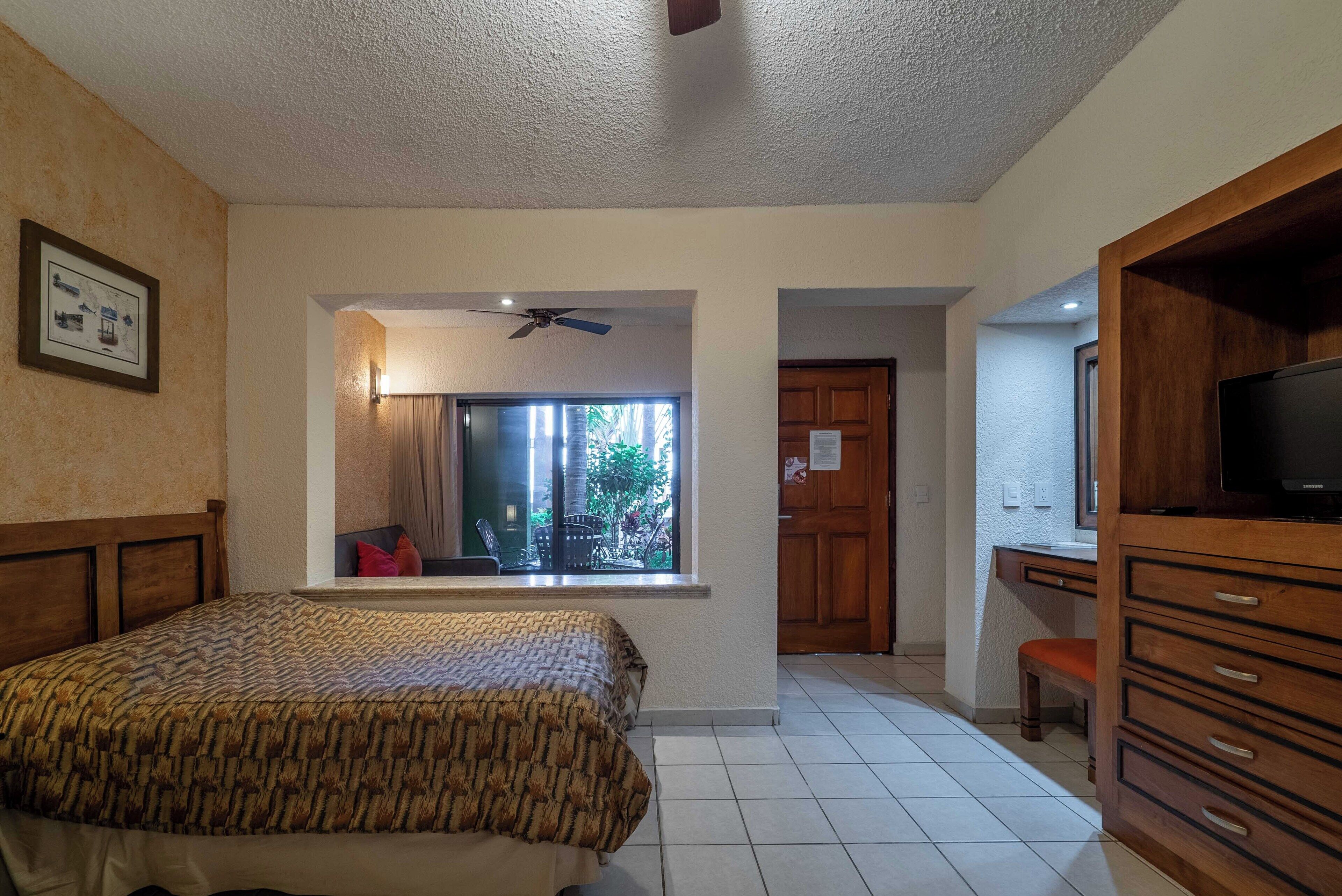 Un dormitorio con cama, escritorio, televisor y ventana con vistas a zonas verdes.