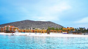 Un hotel frente a la playa con varias edificaciones, una playa de arena y una montaña al fondo.