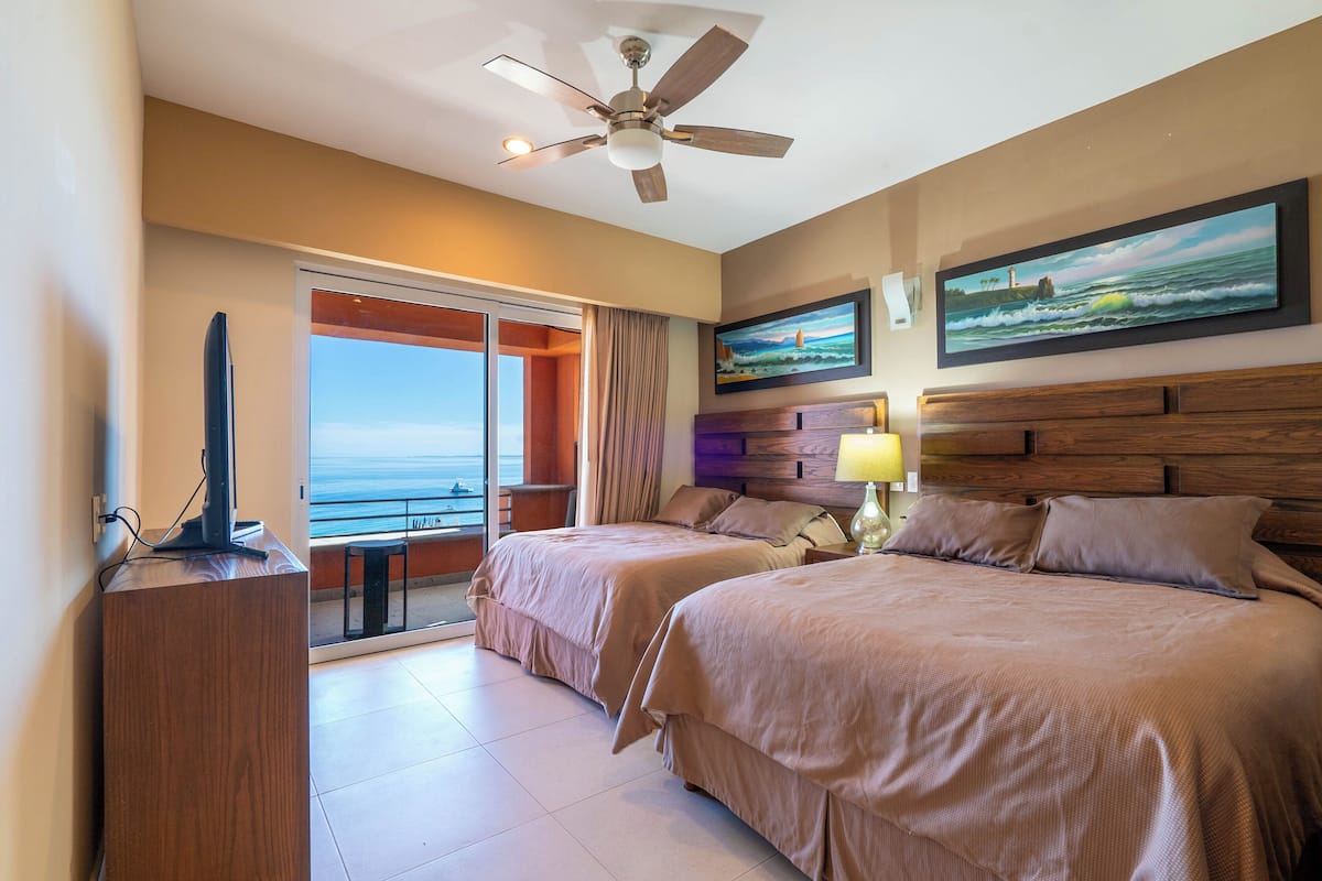 Une chambre d’hôtel avec deux lits, un ventilateur de plafond, une télévision, un tableau et une vue sur l’océan.