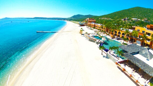 Un complejo turístico en la playa con una extensa playa de arena, mar azul cristalino y una zona de piscina con sombrillas.