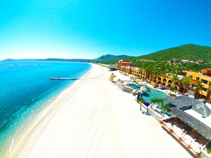 Un complexe hôtelier de bord de mer doté d’une longue plage de sable, d’une mer d’un bleu limpide et d’un espace piscine agrémenté de parasols.