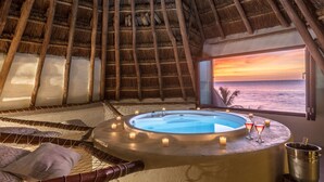 A room with a thatched roof, a circular hot tub with candles, and a view of the sunset over the ocean. - Hotel Villas Flamingos (Isla Holbox)