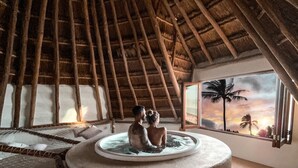 A room with a thatched roof, a circular water feature, and a view of palm trees and a sunset through a window. - Hotel Villas Flamingos (Isla Holbox)