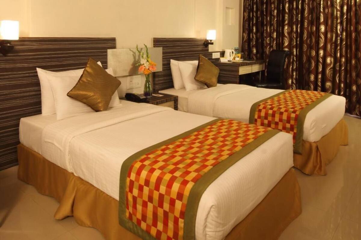 Habitación de hotel con dos camas, cada una con colcha a cuadros, una mesita de noche con lámpara y un panel decorativo en la pared.