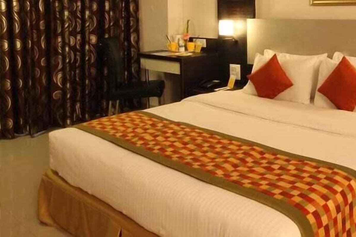 Una habitación de hotel con cama, escritorio, silla y mesita de noche con un jarrón de flores.